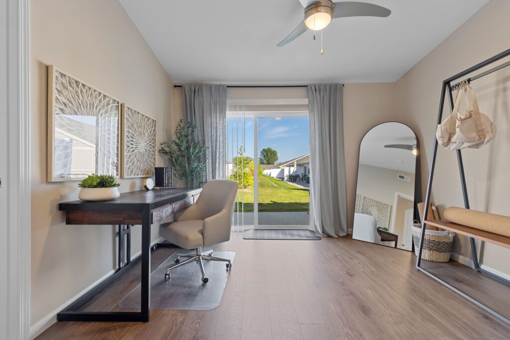 A modern home office with a desk, chair, decor items, a large mirror, bench, clothing rack, and sliding glass doors opening to a grassy backyard.