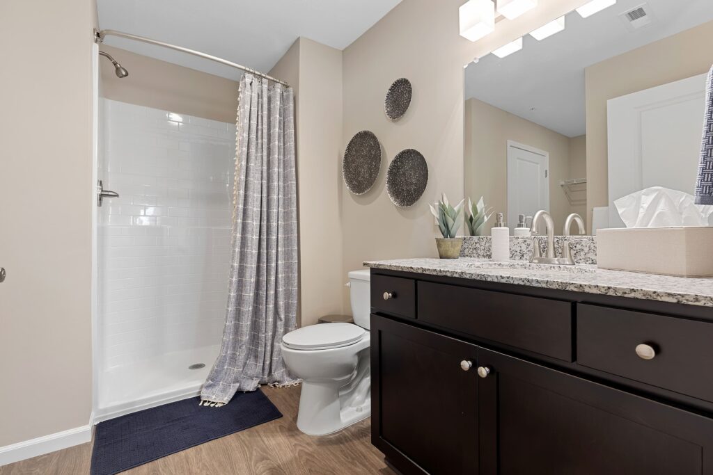 Modern bathroom with a walk-in shower, toilet, dark wood vanity with granite countertop, wall-mounted decor, and neutral-colored walls and flooring.