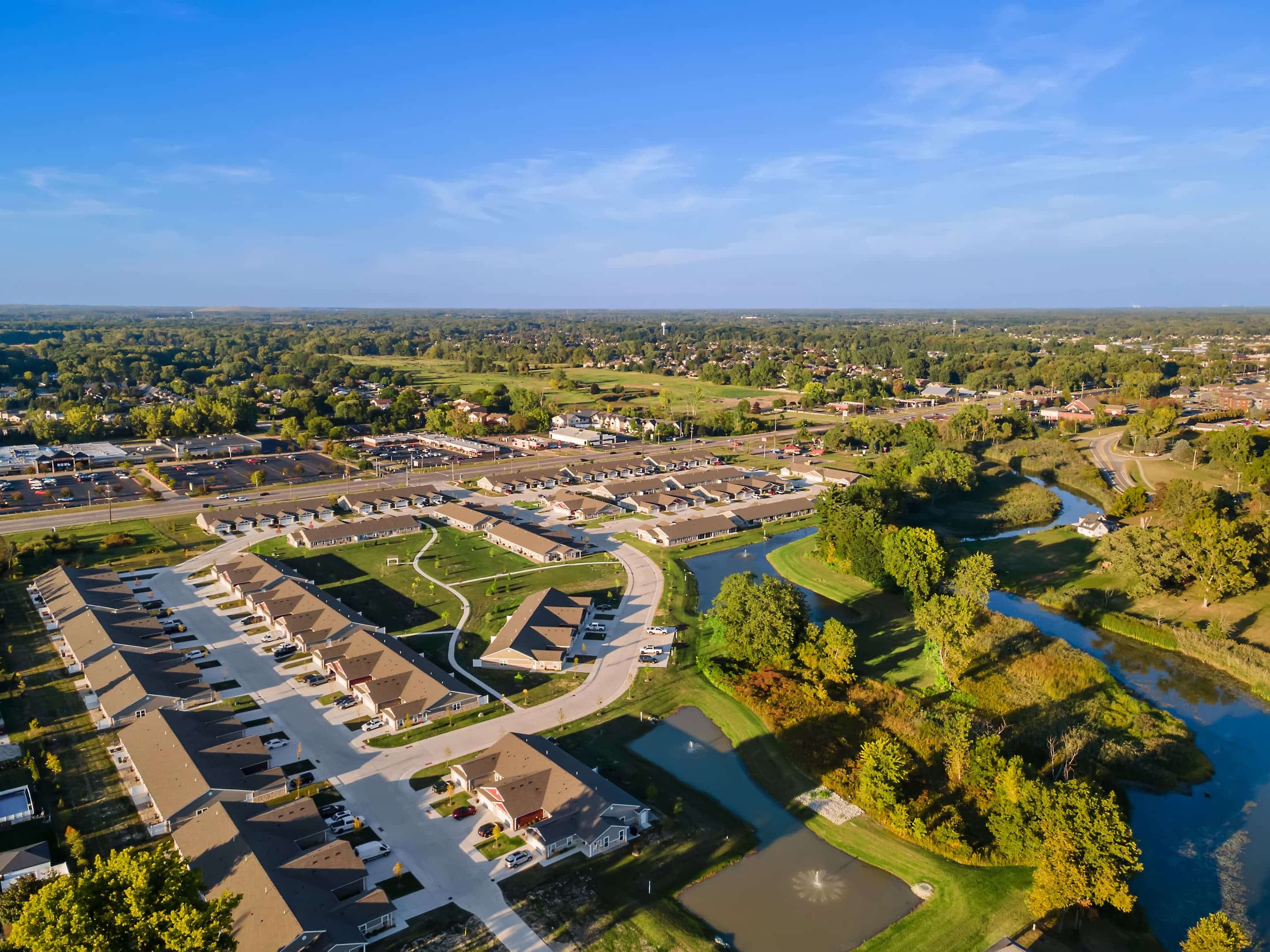 Aerial view of a suburban neighborhood with rows of houses, roads, small ponds, and green trees under a clear blue sky.