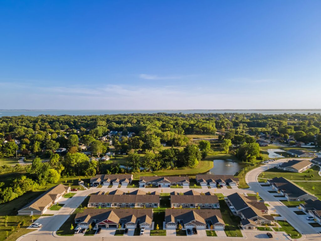 Aerial view of a suburban neighborhood with rows of houses, green lawns, trees, a pond, and a clear blue sky.