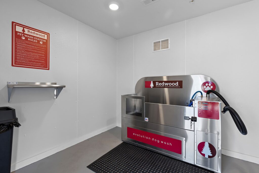 A self-service dog wash station with a metal tub, hose, and dryer in a clean, white room, with a wall sign displaying pet wash rules.