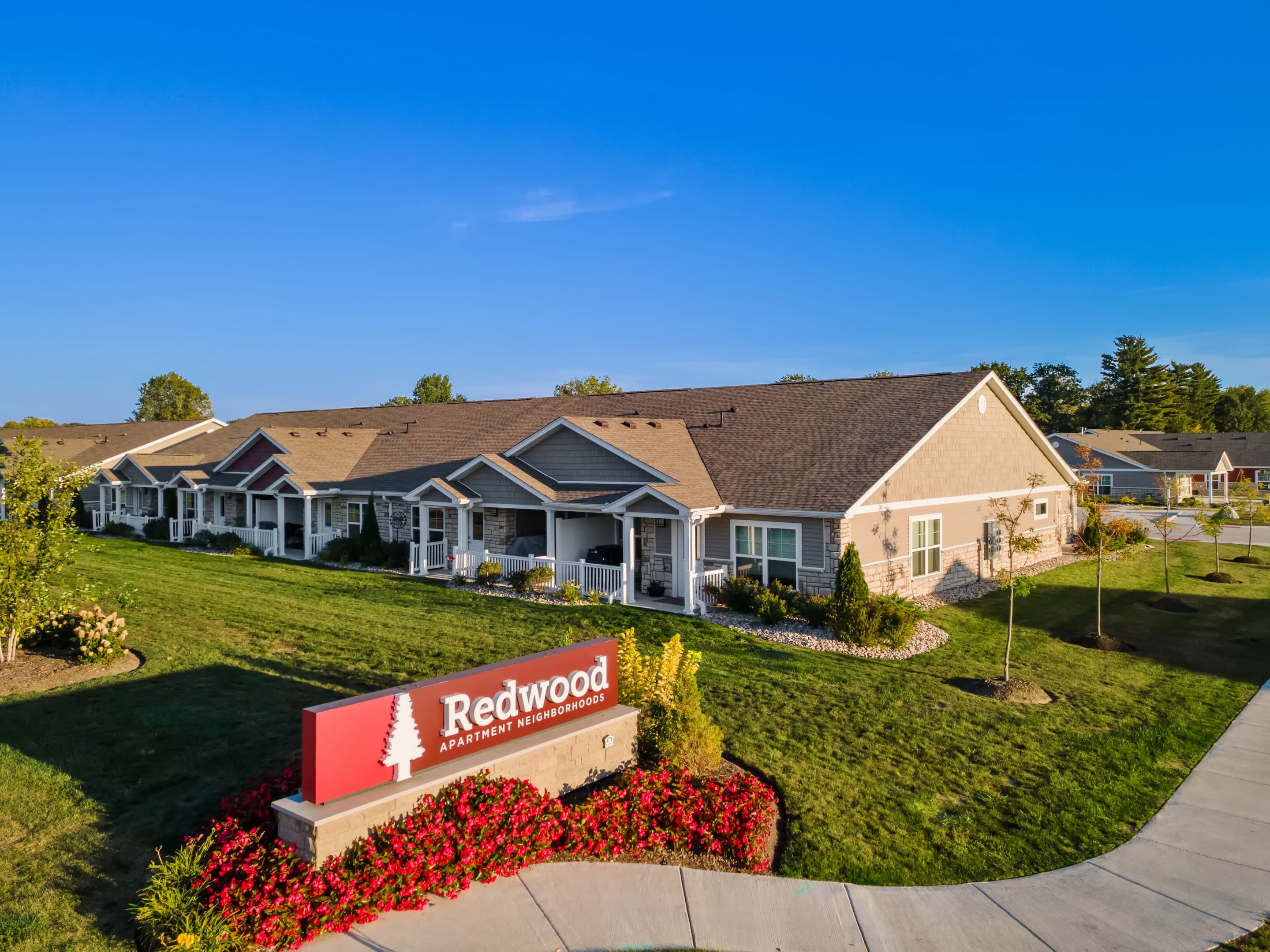 A row of single-story apartment homes with well-kept lawns and landscaping, featuring a red sign that reads "Redwood Apartment Neighborhoods.