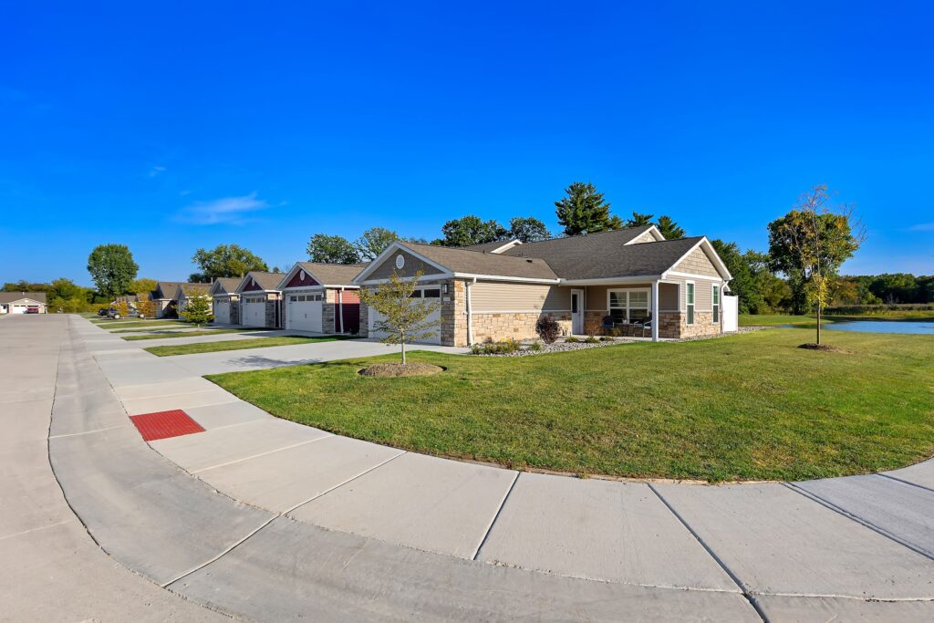 A row of single-story suburban houses with garages, lawns, and a sidewalk under a clear blue sky.