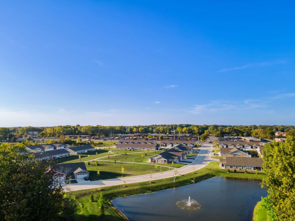 Aerial view of a suburban neighborhood with houses, green lawns, a pond with a fountain, and tree-lined streets under a clear blue sky.