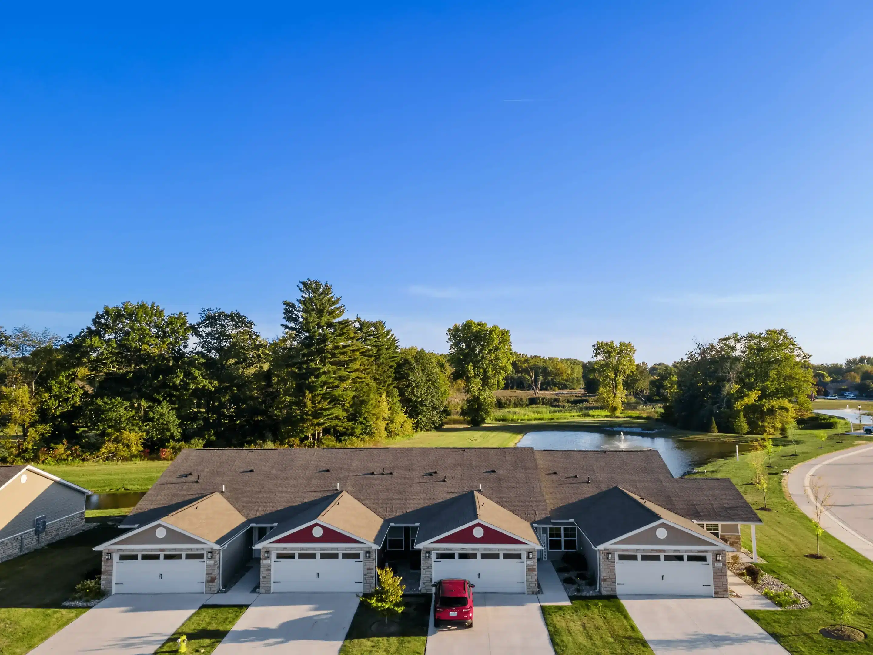 A row of five attached townhouses with garages, one red car parked in a driveway, and a pond with trees in the background under a clear blue sky.