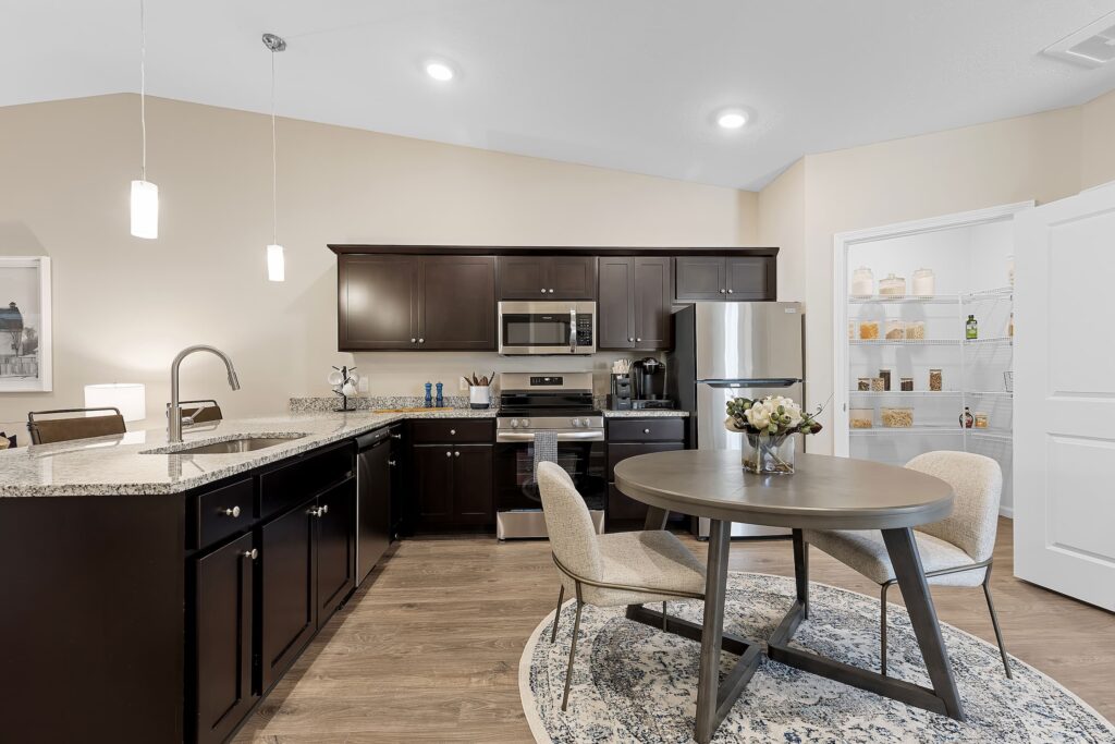Modern kitchen with dark cabinets, stainless steel appliances, a round dining table with two chairs, granite countertops, and an open pantry with shelves.