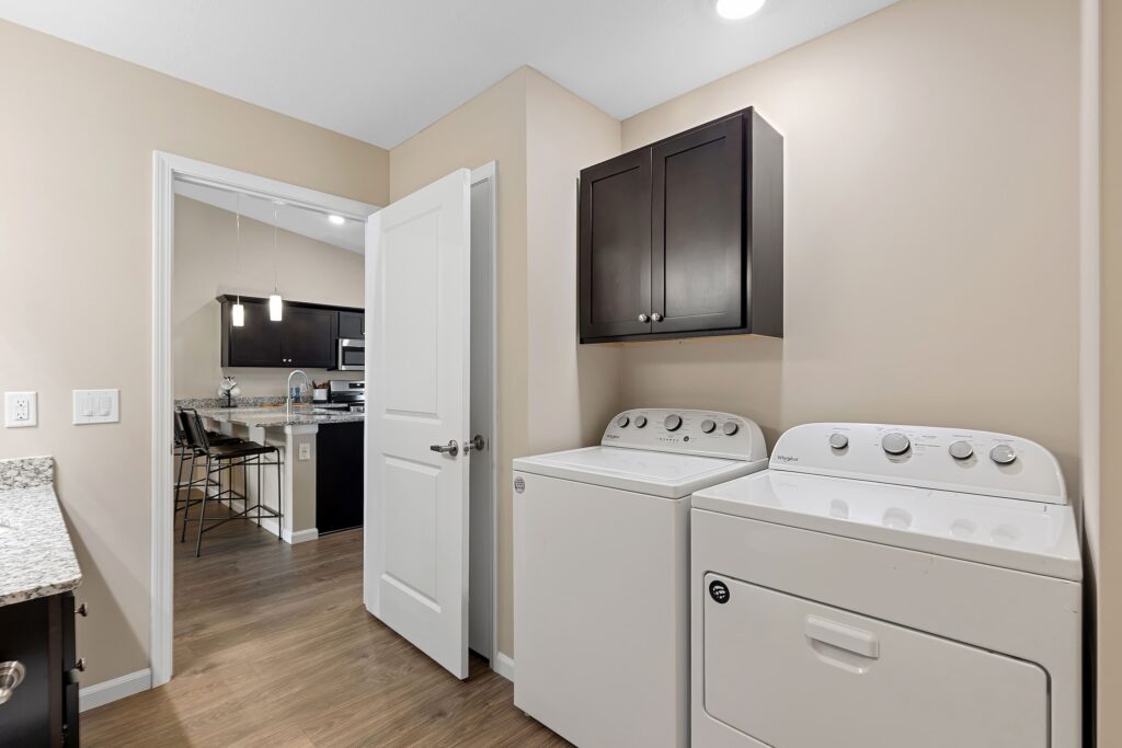 A laundry room with a washing machine and dryer, wood flooring, beige walls, and cabinets; an open door leads to a kitchen area with a dining table and chairs.