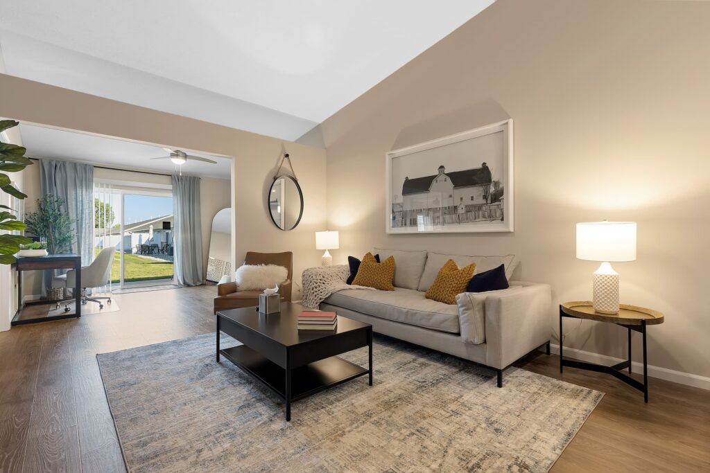 A modern living room with a beige sofa, black coffee table, two side tables with lamps, wall art, and a large window leading to an outdoor patio area.