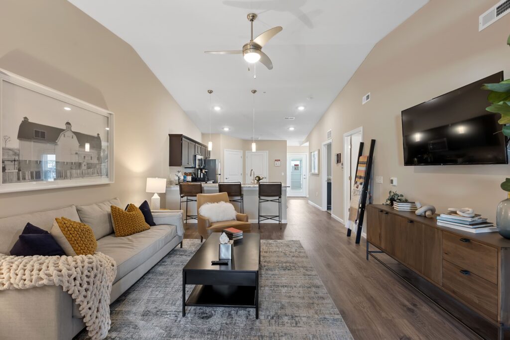 Modern open-concept living room with a beige sofa, dark wood furniture, wall-mounted TV, and kitchen in the background. Ceiling fans and recessed lighting are visible.