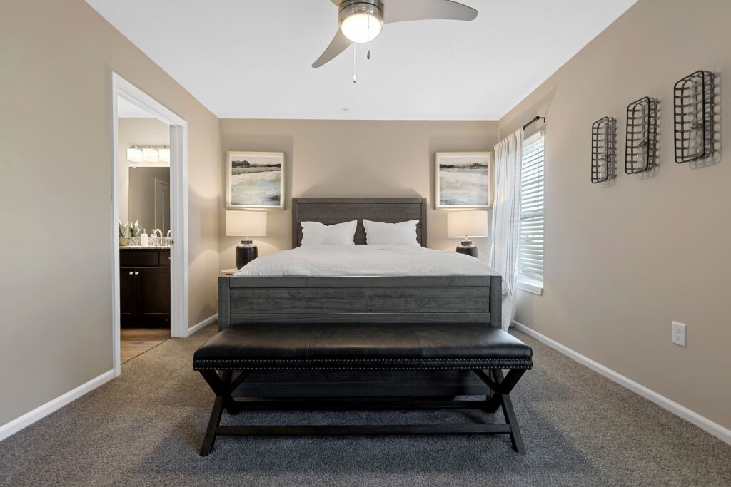 A neatly made bed with two pillows is centered in a bedroom, flanked by nightstands and lamps, with a black bench at the foot and a window with blinds to the right.