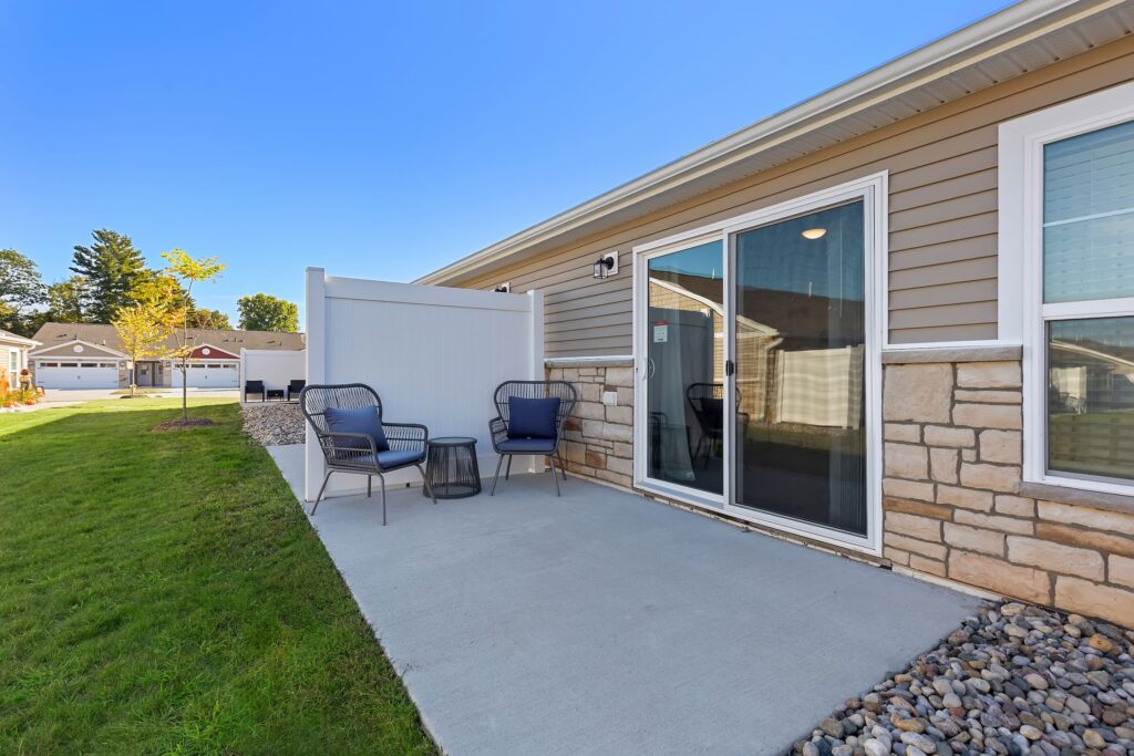 A concrete patio with two chairs, a small table, and a privacy screen beside a single-story house with sliding glass doors and stone siding.