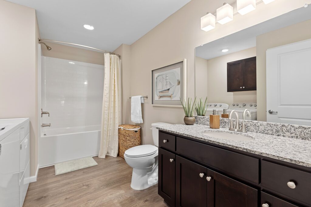 Modern bathroom with wood flooring, dark vanity, granite countertop, large mirror, toilet, laundry machines, and a shower-tub combo with a white curtain and wall art above the toilet.