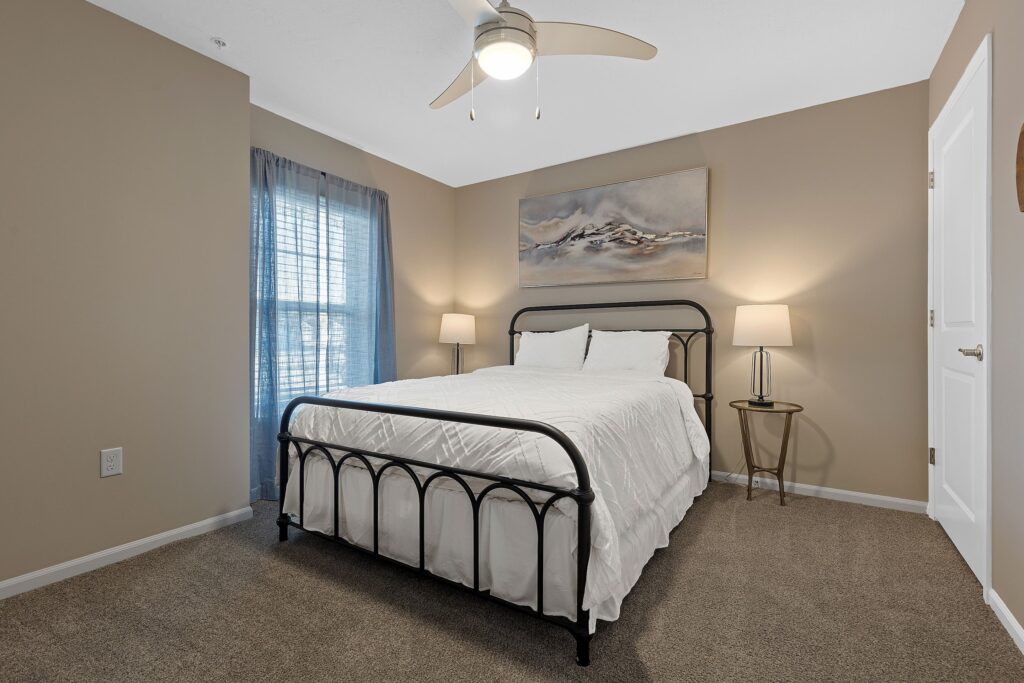 A neatly made bed with a black metal frame in a beige-walled bedroom, flanked by two lamps on either side and a large abstract painting above the headboard.