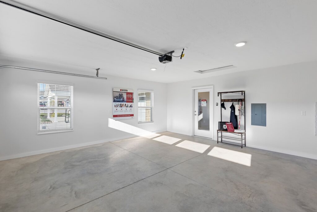 Clean, empty garage interior with concrete floor, white walls, ceiling lights, windows, a door, utility panel, and a metal coat rack with hanging jackets.
