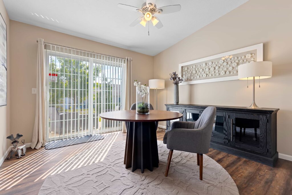 Modern home office with a round table, gray chair, black sideboard, artwork, two lamps, a ceiling fan, and large sliding glass doors letting in natural light.