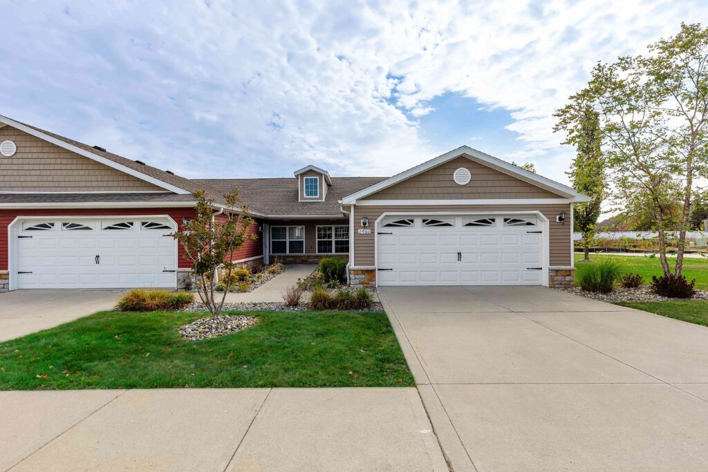 A single-story duplex with two white double garages, beige and red siding, small front lawns, and a driveway under a partly cloudy sky.