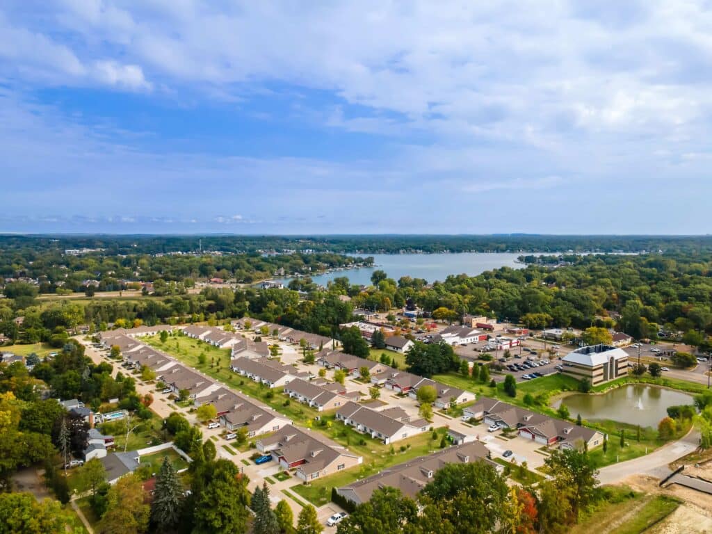 Aerial view of a suburban neighborhood with rows of houses, trees, a lake in the background, and some commercial buildings.