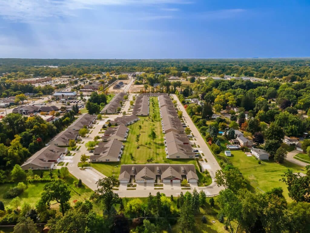 Aerial view of a suburban residential neighborhood with rows of identical houses, green lawns, and surrounding trees under a clear blue sky.