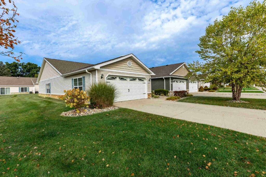 Single-story suburban homes with double garages and well-maintained lawns are shown on a residential street under a partly cloudy sky.