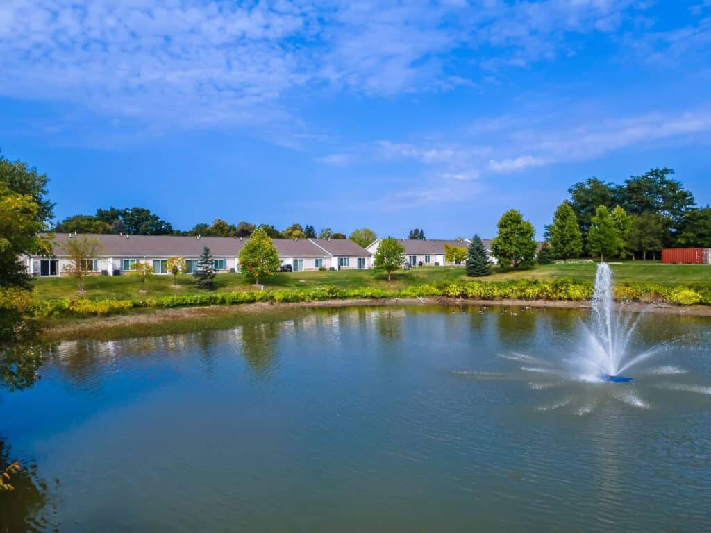 A pond with a fountain in the center, surrounded by green grass, trees, and a row of single-story houses under a blue sky.