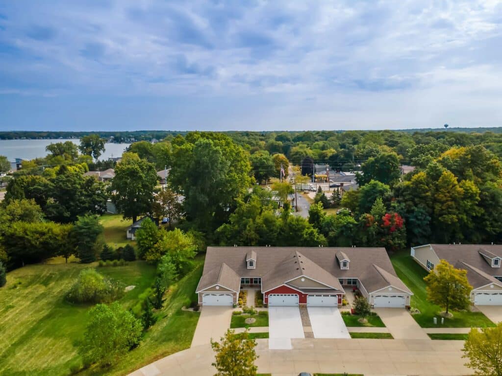 Aerial view of suburban homes with driveways, surrounded by trees and greenery, with a lake and more houses visible in the background under a partly cloudy sky.