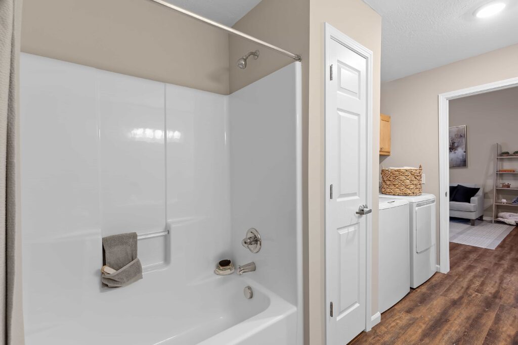 A bathroom with a white bathtub and shower, adjacent to a laundry area with a washer, dryer, and a basket on top, hardwood flooring throughout.