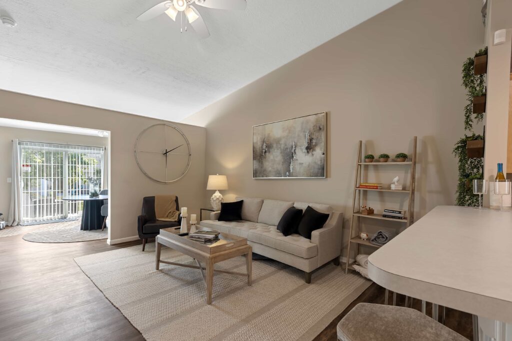 A modern living room with a beige sofa, coffee table, wall art, large clock, open shelves, and a breakfast bar, with natural light coming from a sliding glass door in the background.