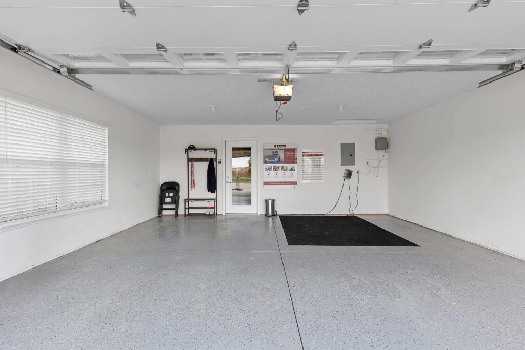 Clean, empty garage with gray floor, a black mat, a coat rack, wall calendar, and an electric panel. Window on the left and closed door at the back.