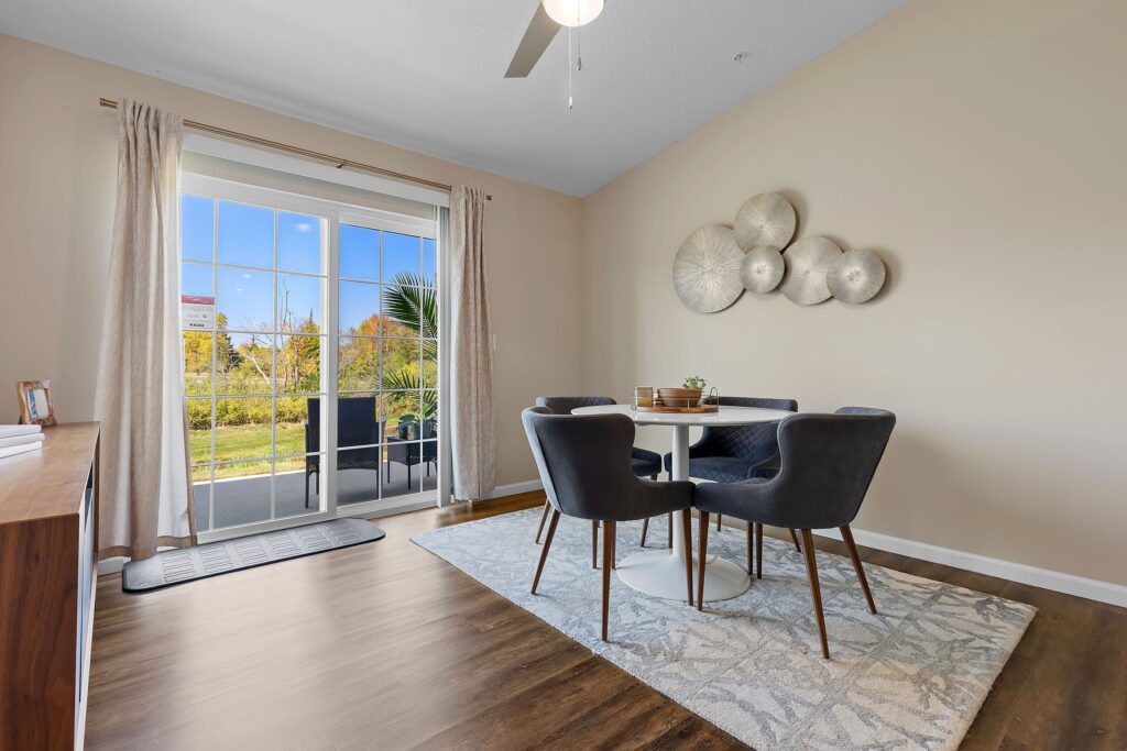 Modern dining area with a round table, four dark chairs, wall art, large window with curtains, and hardwood flooring, overlooking a green outdoor space.