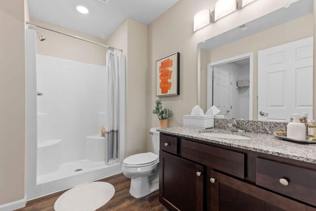 Modern bathroom with a walk-in shower, white curtain, toilet, dark wood vanity with granite countertop, and wall art above a tissue box.