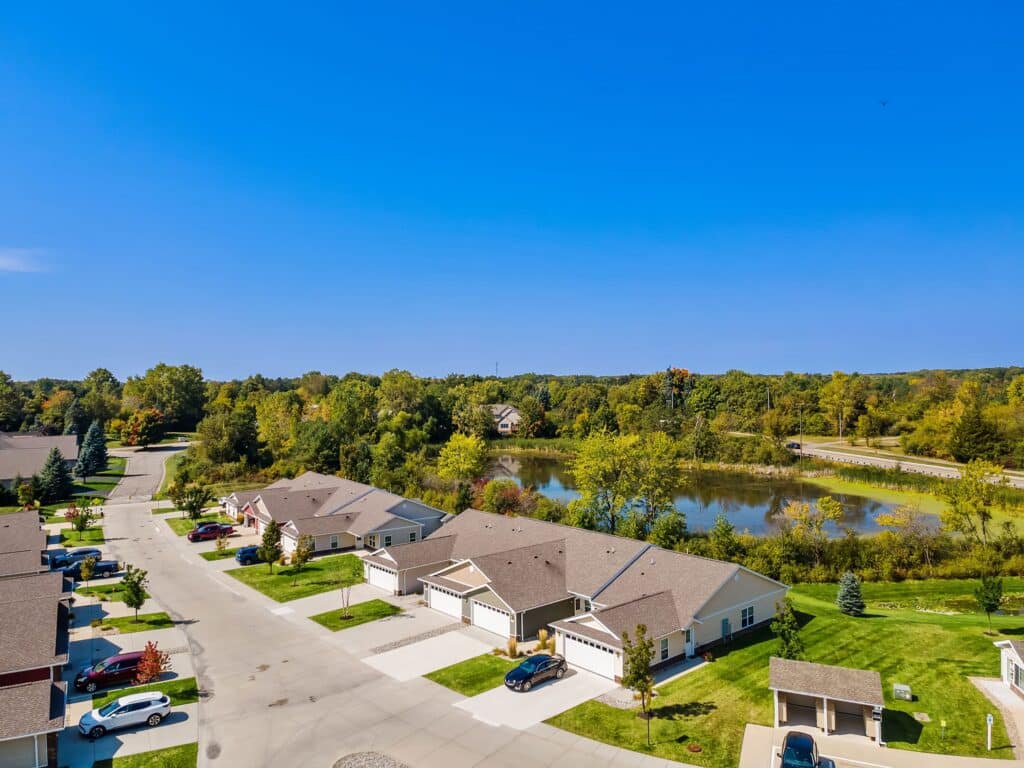Aerial view of a suburban neighborhood with single-story houses, a curved street, and a pond surrounded by trees under a clear blue sky.