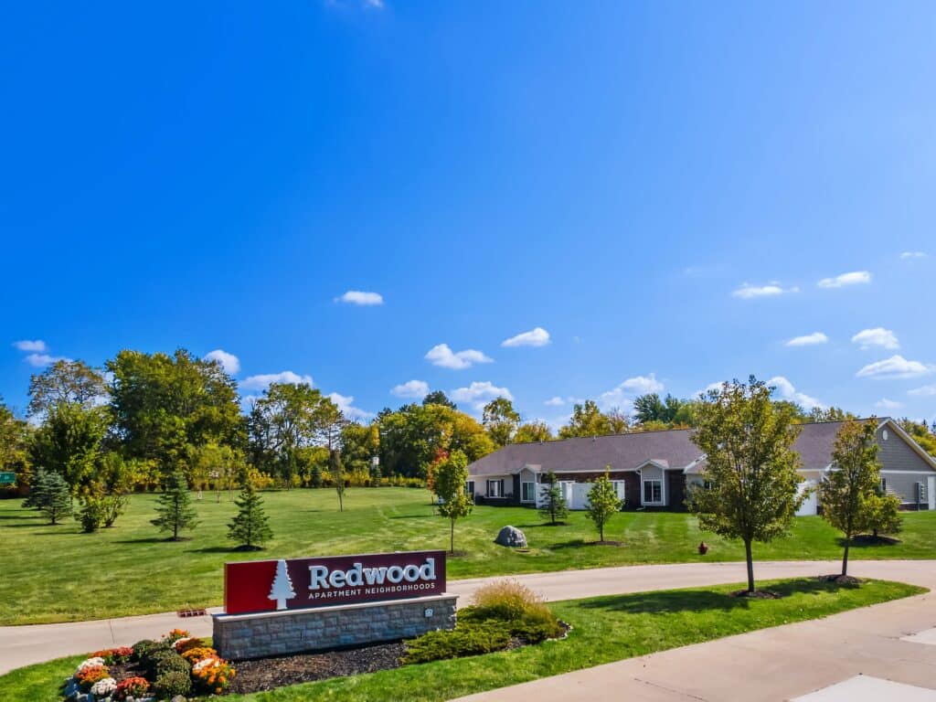 A single-story apartment building sits behind a landscaped lawn and a sign reading "Redwood Apartment Neighborhoods" under a clear blue sky.
