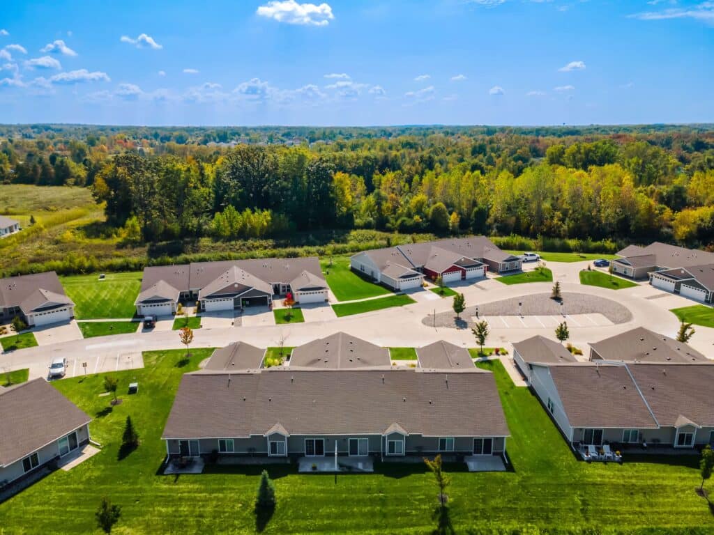 Aerial view of a suburban neighborhood with single-story houses, green lawns, a cul-de-sac, and trees in the background under a blue sky.