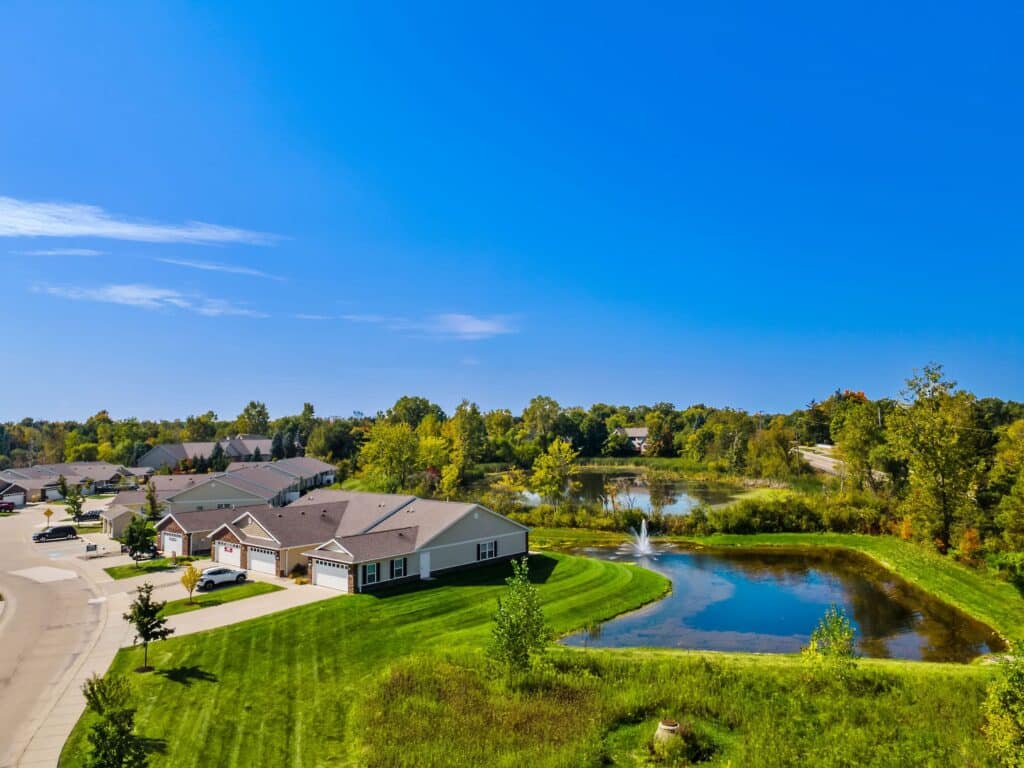 A suburban neighborhood with single-story houses, a small pond with a fountain, green lawns, and trees under a clear blue sky.