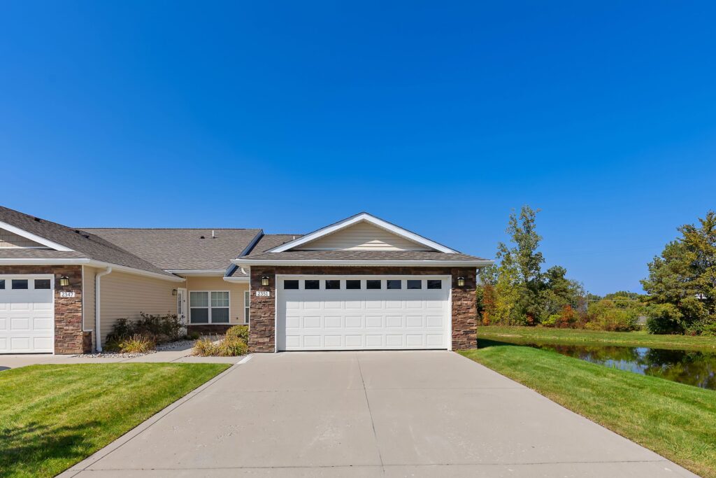 Single-story suburban house with a two-car garage, brick and siding exterior, concrete driveway, and a grassy lawn under a clear blue sky.