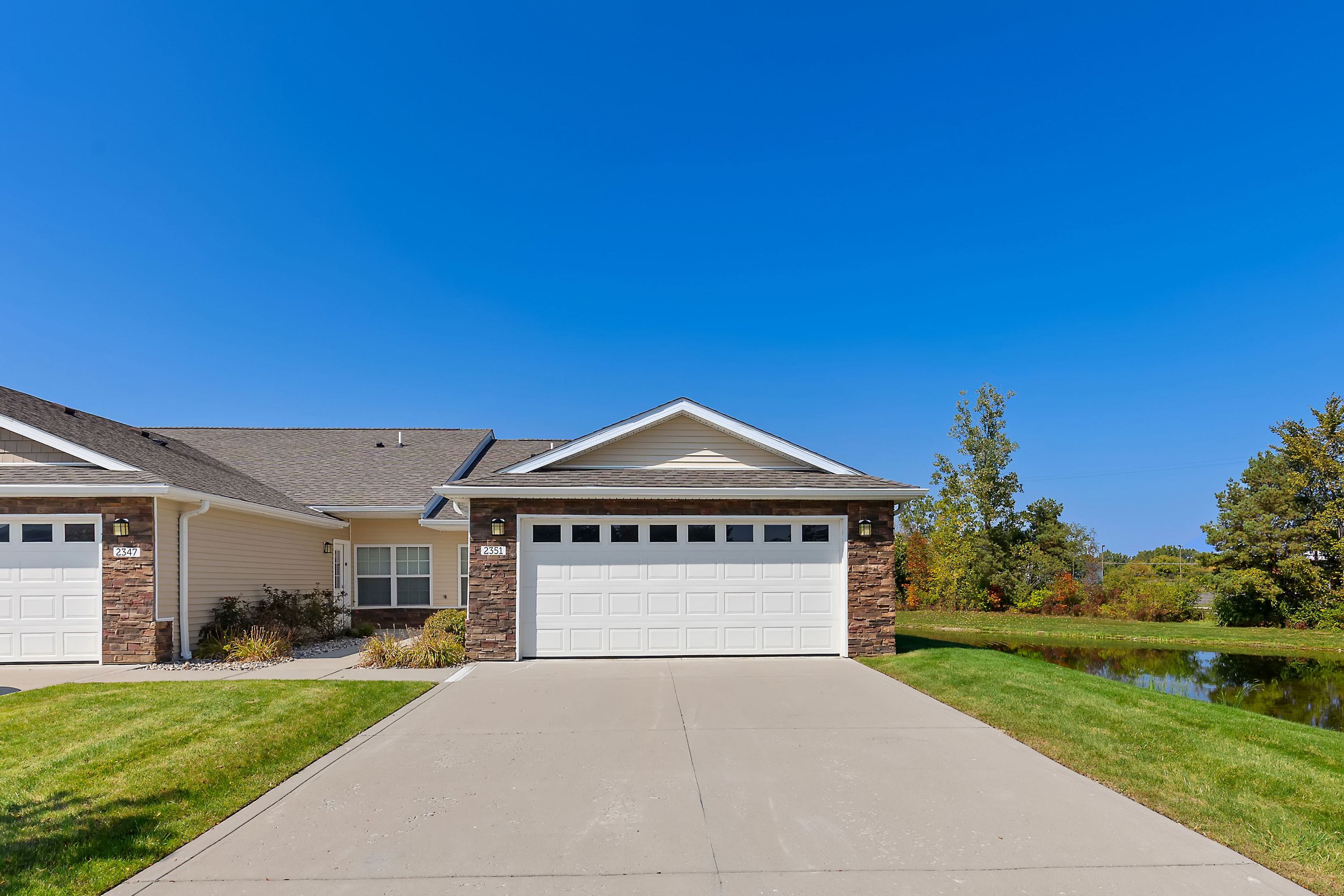 Single-story suburban house with a two-car garage, brick and siding exterior, concrete driveway, and a grassy lawn under a clear blue sky.
