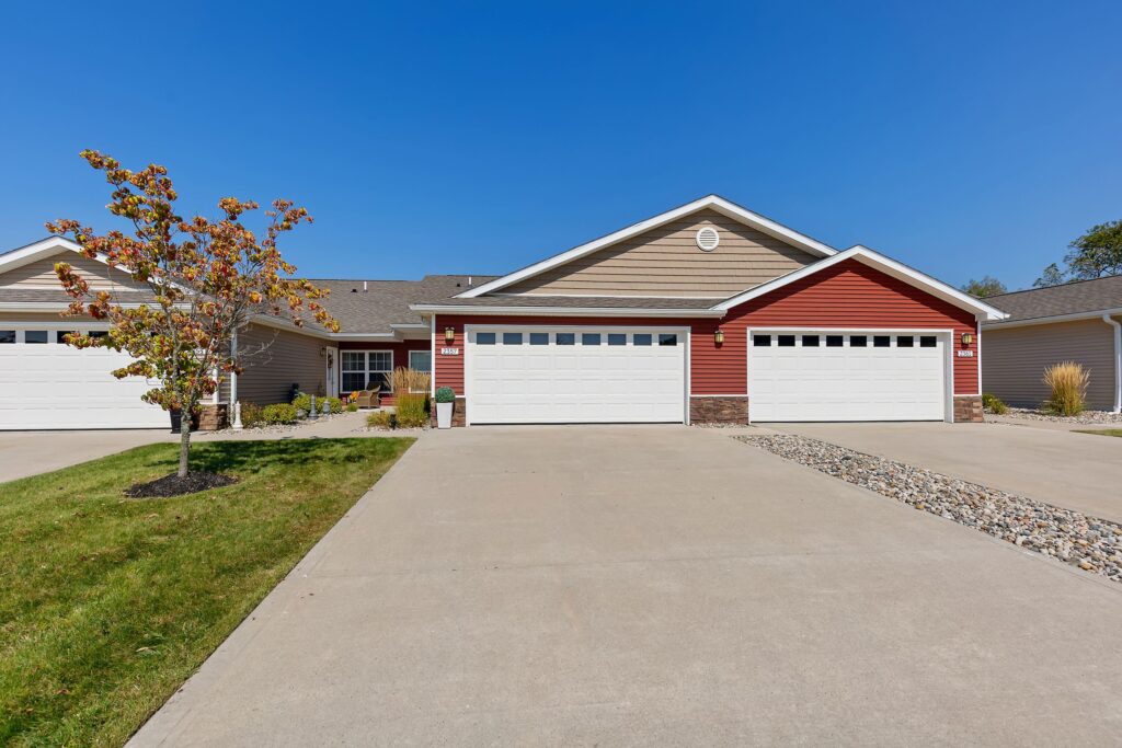 A suburban house with red and tan siding, two white garage doors, a wide driveway, and a small tree in the front yard under a clear blue sky.
