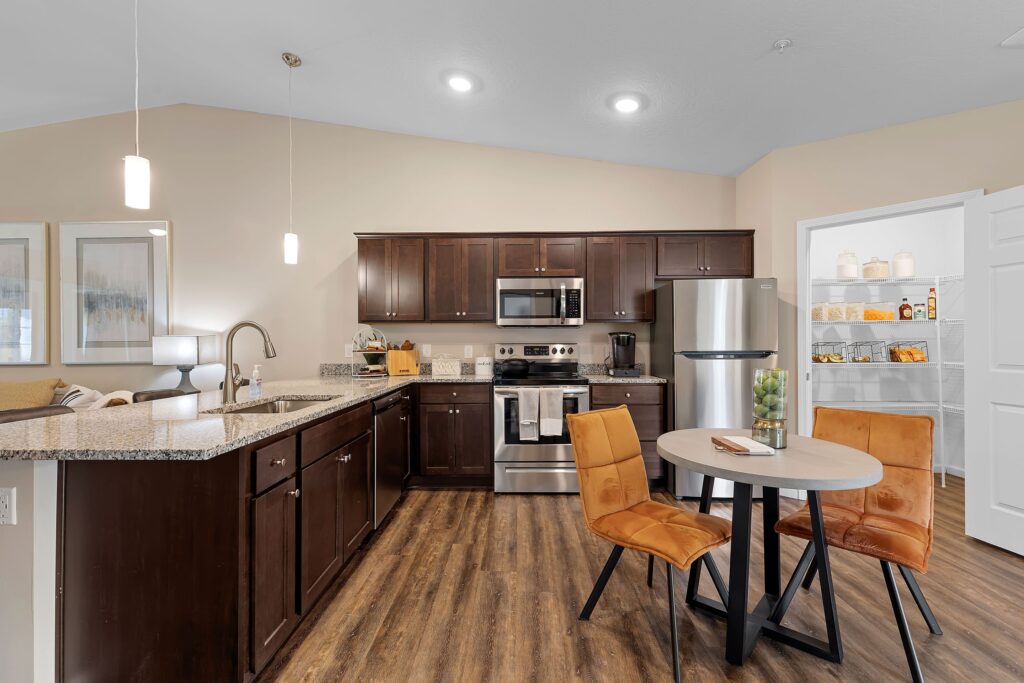 Modern kitchen with dark wood cabinets, stainless steel appliances, a round table with two orange chairs, and an open pantry door.