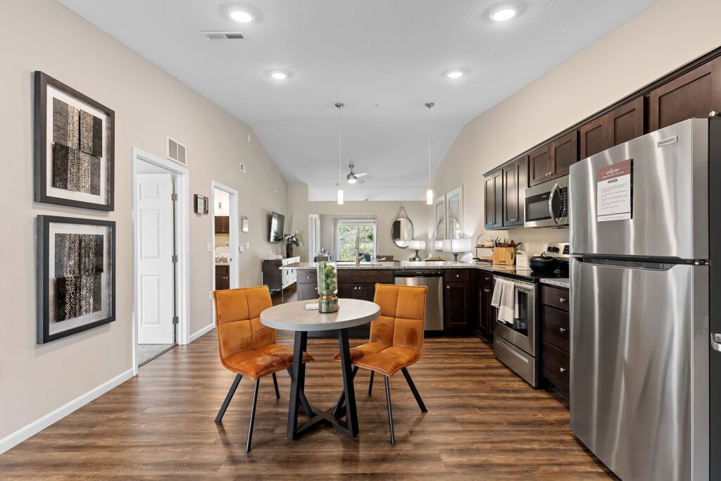Modern apartment kitchen with stainless steel appliances, dark wood cabinets, a small round dining table, and two orange chairs on wood flooring.