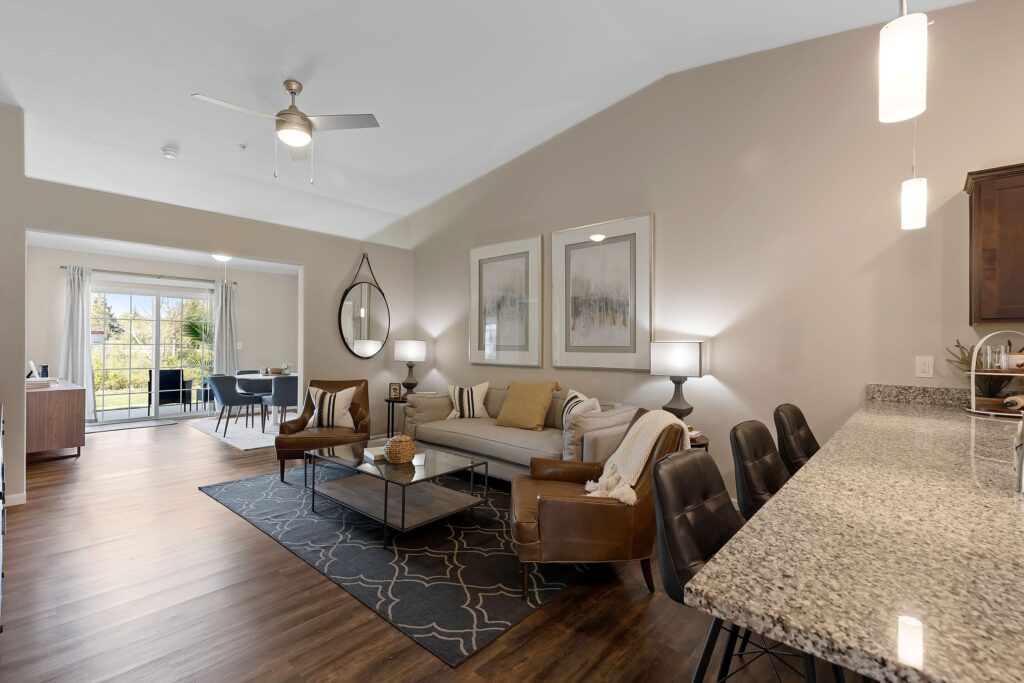 Modern open-concept living room and kitchen with granite countertops, neutral decor, leather chairs, large windows, and dining area in the background.