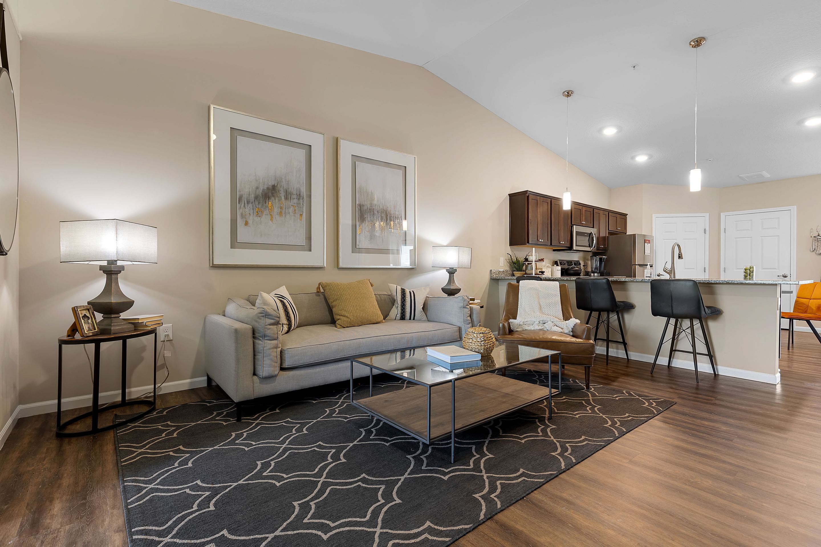 Modern living room with a gray sofa, two framed artworks, lamps, a patterned rug, coffee table, and an open kitchen with bar stools in the background.
