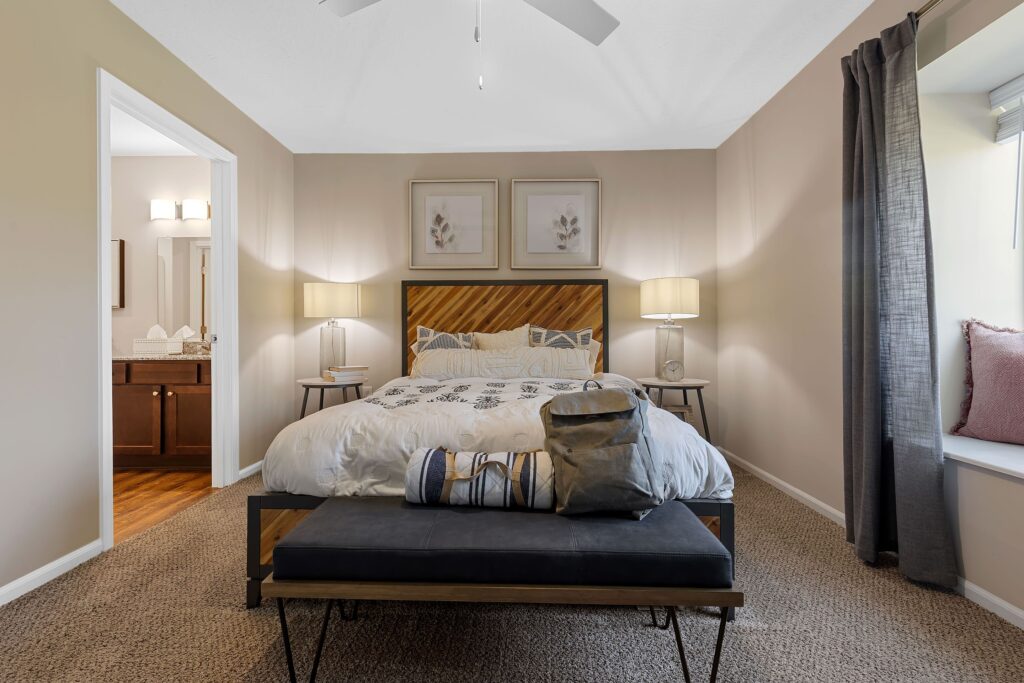 A neatly made bed with pillows and a bench at its foot is centered in a bedroom. Two lamps, framed artwork, and a window seat with cushions are also visible.
