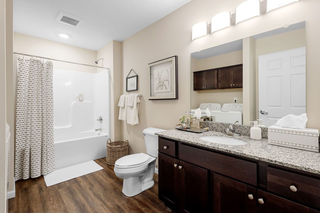 Modern bathroom with a bathtub and shower, toilet, granite countertop with sink, dark wood cabinets, washer and dryer, and neutral decor.