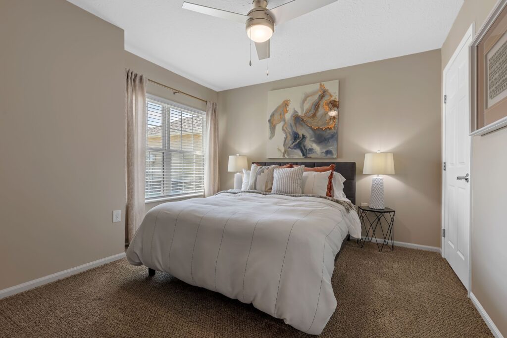 A neatly made bed with white bedding in a small bedroom, flanked by two lamps, with a ceiling fan above and a window allowing natural light in.