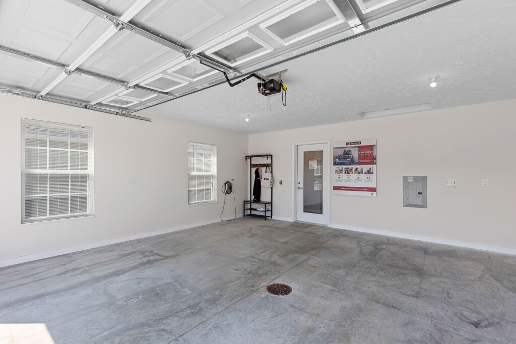 Clean, empty garage with concrete floor, open automatic garage door, two windows, a glass door, and minimal wall-mounted utility features.