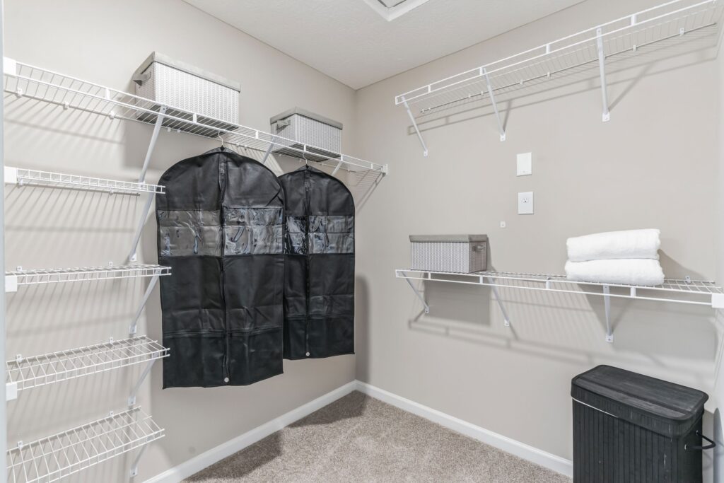 A walk-in closet with wire shelving, two black garment bags, gray storage boxes, two folded white towels, and a black bin on carpeted flooring.