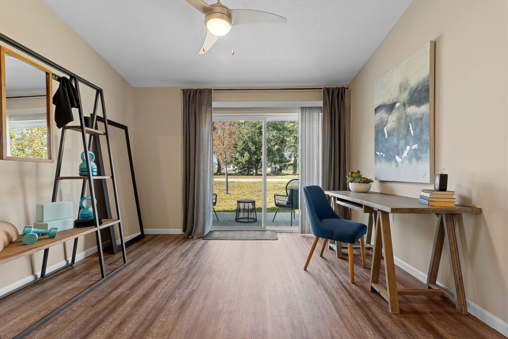 A home office features a wooden desk with a blue chair, a shelving unit with yoga equipment, large window doors, and a view of an outdoor patio and trees.
