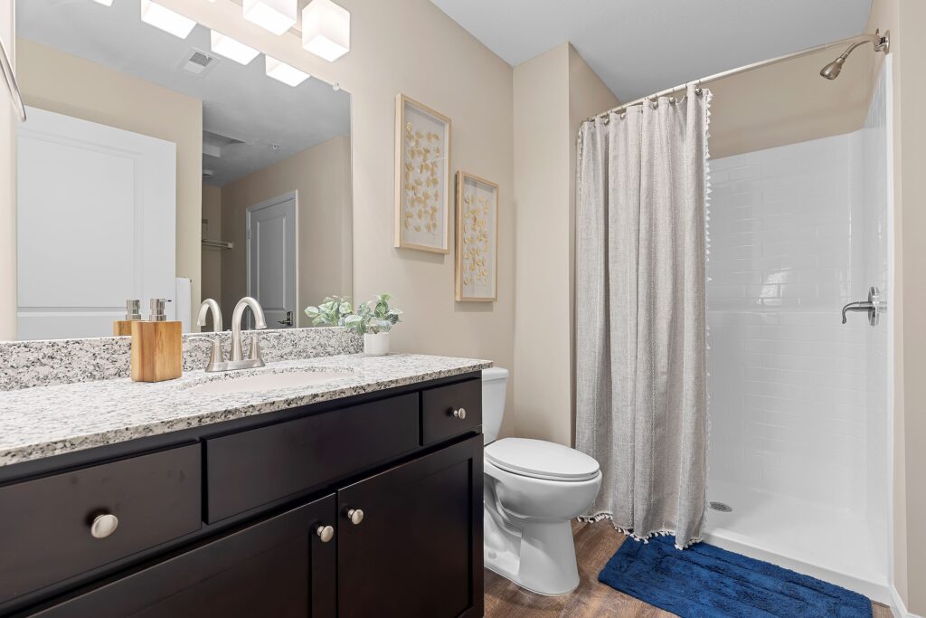 Modern bathroom with granite countertop, dark wood vanity, toilet, walk-in shower with curtain, blue rug, and beige walls with framed artwork.