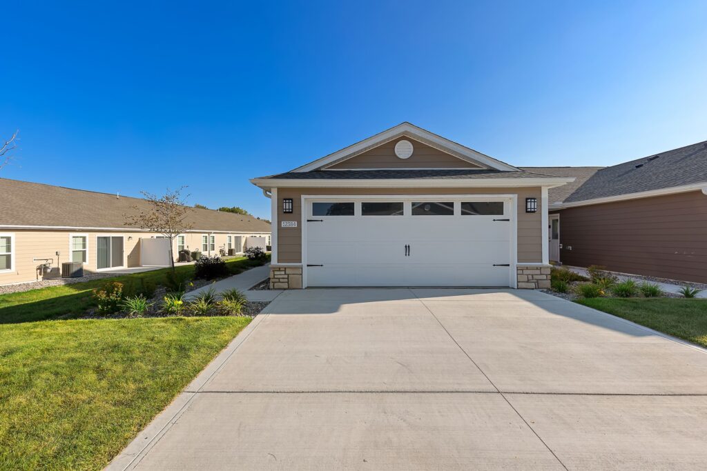 Single-story house with a double garage, light brown siding, stone accents, and a clean concrete driveway under a clear blue sky.