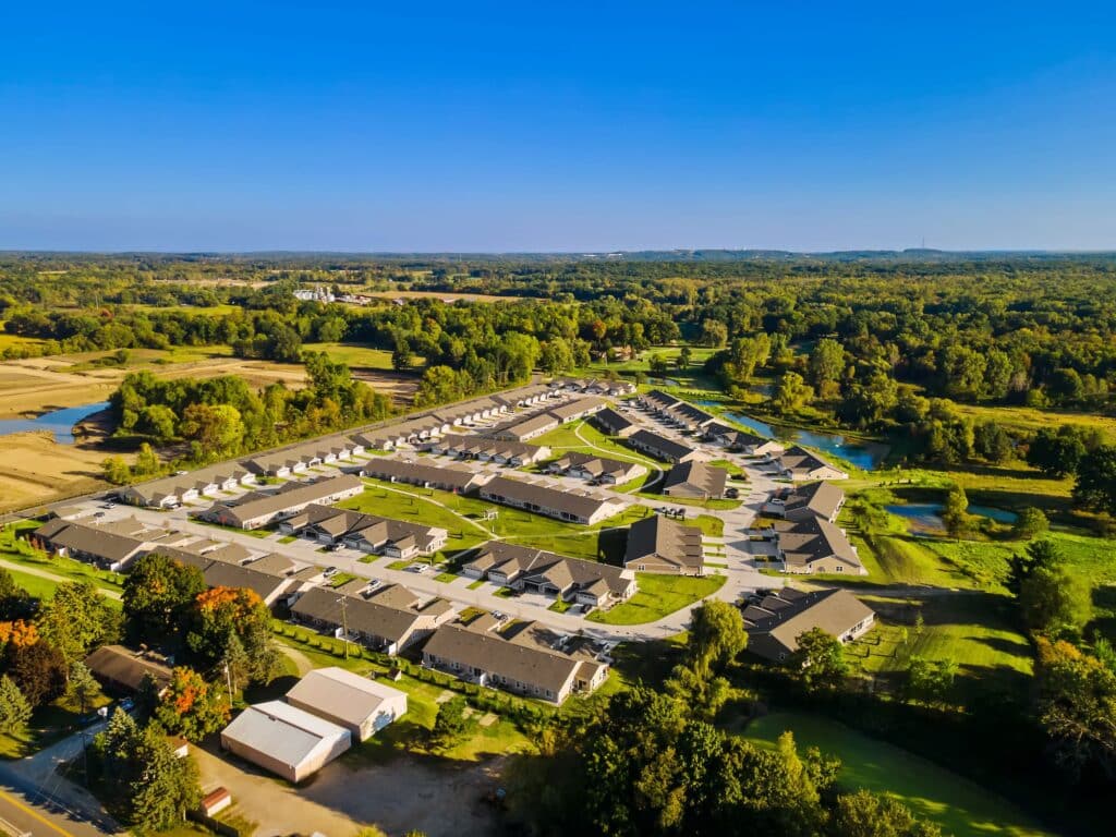 Aerial view of a suburban residential neighborhood with rows of houses, green lawns, surrounding trees, and a few ponds under a clear blue sky.
